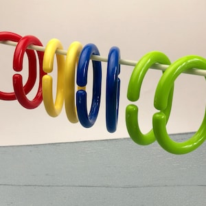 Può includere: Sei anelli per tenda da doccia in plastica colorati appesi a un'asta da doccia bianca. Gli anelli sono rosso, giallo, blu, verde e verde.