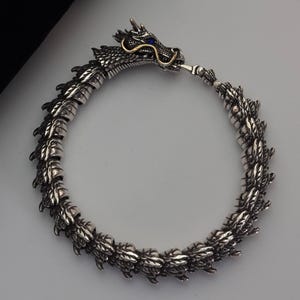 Puede incluir: Pulsera de dragón plateada con detalles intrincados. La cabeza del dragón presenta detalles dorados y un ojo de gema azul. La pulsera está hecha de segmentos unidos.