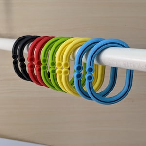 Könnte beinhalten: Ein Satz von neun bunten Silikon-Duschvorhanghaken in Schwarz, Rot, Grün, Gelb und Blau. Die Haken hängen an einer weißen Duschstange.