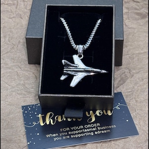 Peut inclure: Un collier pendentif avion argenté présenté dans un coffret cadeau noir. Le collier présente un design d'avion détaillé suspendu à une chaîne. Une carte de remerciement avec des lettres dorées est visible.