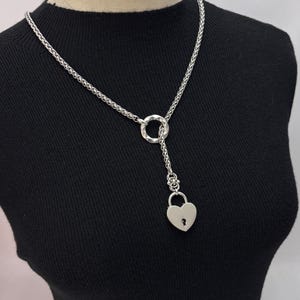 Puede incluir: Collar plateado con un colgante de candado en forma de corazón. La cadena tiene una textura y un acento circular. El colgante cuelga de una cadena delicada, creando un accesorio elegante y único.