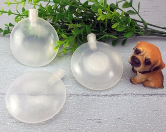 Op de afbeelding: Drie transparante, piepende hondenspeelgoedjes van plastic met een klein handvat aan de bovenkant. De speelgoedjes liggen op een lichtgrijze houten ondergrond. Een klein bruin hondje staat op de achtergrond.