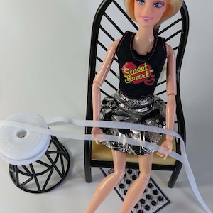 May include: A doll wearing a black tank top with a red heart design and a silver skirt, seated in a black chair. A roll of white ribbon and a small black button card are nearby. The doll is wearing pink shoes.