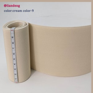 Peut inclure: Un rouleau de bande &eacute;lastique de couleur cr&egrave;me, mesurant 23 cm de long. La bande &eacute;lastique est enroul&eacute;e et un m&egrave;tre ruban est visible &agrave; c&ocirc;t&eacute;. Le texte "color:cream color-9" et "@liandeng" sont visibles dans le coin sup&eacute;rieur gauche de l'image.