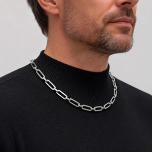 May include: A silver-toned chain necklace featuring elongated, rectangular links with rounded edges. The necklace is worn around the neck, resting against a black ribbed mock neck shirt, showcasing a clean, metallic finish and a uniform link design.