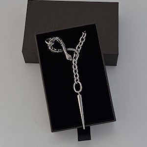 Op de afbeelding: Een zilverkleurige ketting met een hanger in de vorm van een kronkelende slang, verbonden aan een dikke ketting die uitloopt in een lange, puntige bedel. Het sieraad wordt gepresenteerd in een zwarte rechthoekige doos met fluwelen voering.