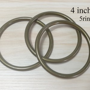 Puede incluir: Tres anillos metálicos de color bronce, cada uno de aproximadamente 10 cm de diámetro, están entrelazados. El texto "4 inch 5 rings" es visible en la esquina superior derecha. Los anillos están sobre una superficie de madera clara.