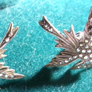 Puede incluir: Primer plano de dos pendientes plateados con forma de pájaro. Cada pendiente presenta un diseño de ala detallado con cristales transparentes incrustados. Los pendientes están colocados sobre un fondo turquesa, resaltando sus intrincados detalles y acentos brillantes.