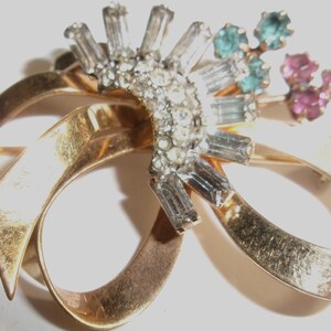 Peut inclure: Une broche en métal doré en forme de nœud ruban, ornée d'un groupe central de strass transparents taille baguette et ronds disposés en éventail, agrémentée de petits groupes de pierres rondes bleu clair et roses sur le côté supérieur droit.