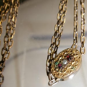 May include: A gold-toned metal chain necklace featuring an oval-shaped pendant with intricate filigree detailing. The center of the pendant contains a rectangular inlay set with two small dark red stones and one central white stone in a vertical row.