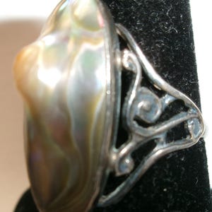 Könnte beinhalten: Ein Ring mit einem großen, ovalen, irisierenden Abalone-Muschel-Cabochon in wirbelnden Grün-, Braun- und Perlmutttönen. Die Fassung besteht aus silberfarbenem Metall mit einem durchbrochenen, schnörkelartigen Design an den Seiten.