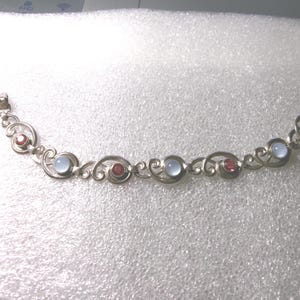 Könnte beinhalten: Ein silberfarbenes Metallarmband mit einer Reihe von miteinander verbundenen, geschwungenen Gliedern. Das Design wechselt zwischen runden, durchscheinenden hellblauen Cabochon-Steinen und kleineren, runden, facettierten dunkelroten Steinen.