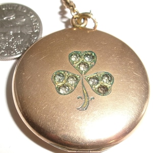 May include: A round gold-tone metal locket featuring a central shamrock design inlaid with small clear stones. The locket is attached to a gold-tone chain and is positioned next to a United States dime for scale, showing the locket is slightly larger.
