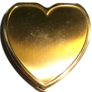 May include: A heart-shaped, gold-colored compact mirror. The compact has a smooth, reflective surface with a slight sheen. The edges are rounded and the compact appears to be made of metal. The compact is closed and has a clasp.