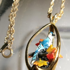 May include: Gold-coloured necklace with a pendant depicting a blue jay on a branch with autumn leaves. The bird is set within a teardrop-shaped frame. The chain is a rope style.