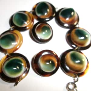May include: A bracelet composed of circular links featuring polished, dome-shaped centres with swirling patterns of deep green, amber, and cream colours. The links are connected by small metal rings, and the piece includes a silver-toned metal clasp.
