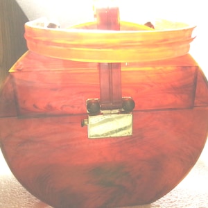 May include: A round handbag crafted from translucent amber-colored material with a marbled pattern. The bag features a semi-circular handle attached to the top and a rectangular metallic clasp centered on the front, securing the two halves together.