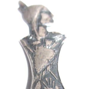 Könnte beinhalten: Ein silberfarbener Metallanhänger in Form einer stilisierten Figur, möglicherweise einer Frau oder eines Fabelwesens. Der Anhänger hat ein detailliertes Design mit strukturierter Oberfläche und einem fließenden, drapierten Gewand. Der Kopf der Figur ist nach oben geneigt.