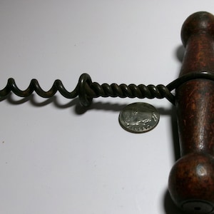 Könnte beinhalten: Ein antiker Korkenzieher mit einem dunkelbraunen, gedrechselten T-förmigen Holzgriff und einem langen, gedrehten Metallschaft, der in einer scharfen, spiralförmigen Schnecke endet. Eine US-Münze liegt zur Größenorientierung neben dem Schaft.