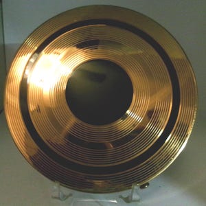 Op de afbeelding: Een goudkleurig, cirkelvormig object met concentrische groeven en een donkere centrale cirkel. Het object heeft een glanzend, reflecterend oppervlak, waarschijnlijk van metaal. Twee donkere banden omcirkelen het midden.