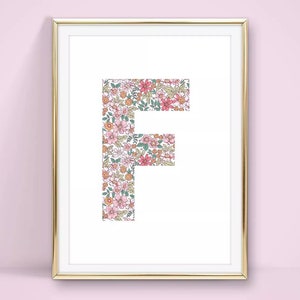May include: A framed print displaying the letter 'F' filled with a floral pattern. The design incorporates pink, orange, and green flowers. The print is encased in a gold frame, suitable for wall decor, adding a touch of elegance.