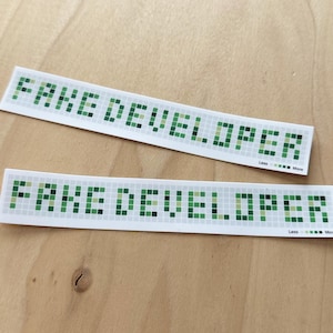 May include: Two white stickers with the text "FAKE DEVELOPER" in a pixelated green font. The stickers are rectangular and slightly curved, with a small graphic and the words "Less ... More" at the bottom.