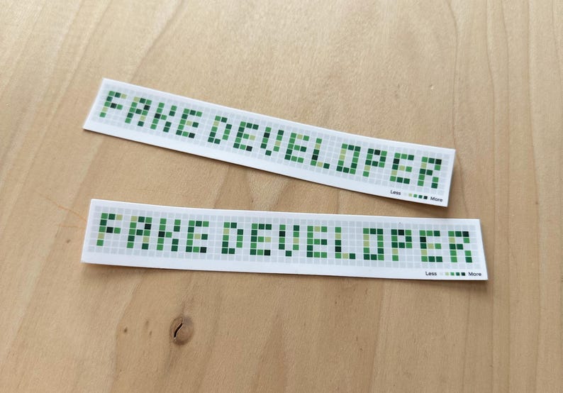 May include: Two white stickers with the text "FAKE DEVELOPER" in a pixelated green font. The stickers are rectangular and slightly curved, with a small graphic and the words "Less ... More" at the bottom.