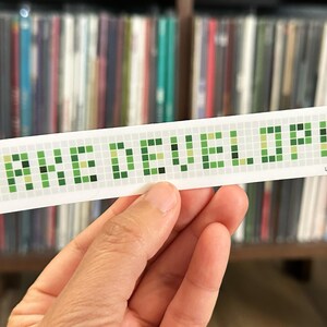 May include: A white sticker with the text "FAKE DEVELOPER" in green pixelated letters. The sticker also has the text "Less More" at the bottom. The sticker is held in front of a shelf of vinyl records.