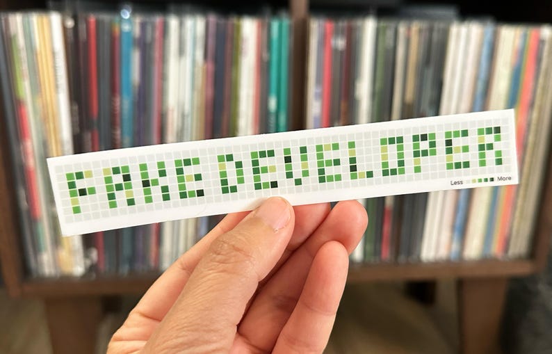 May include: A white sticker with the text "FAKE DEVELOPER" in green pixelated letters. The sticker also has the text "Less More" at the bottom. The sticker is held in front of a shelf of vinyl records.