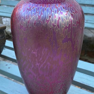 Puede incluir: Un jarrón de vidrio bulboso con un color base magenta intenso y una capa iridiscente de oro metálico. La superficie muestra un patrón orgánico repetitivo de formas alargadas y redondeadas que brillan con un acabado reflectante.