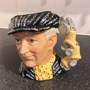 May include: A ceramic mug shaped like a man's head wearing a black and white patterned cap. The mug has a yellow and red polka dot scarf and a handle shaped like a car.