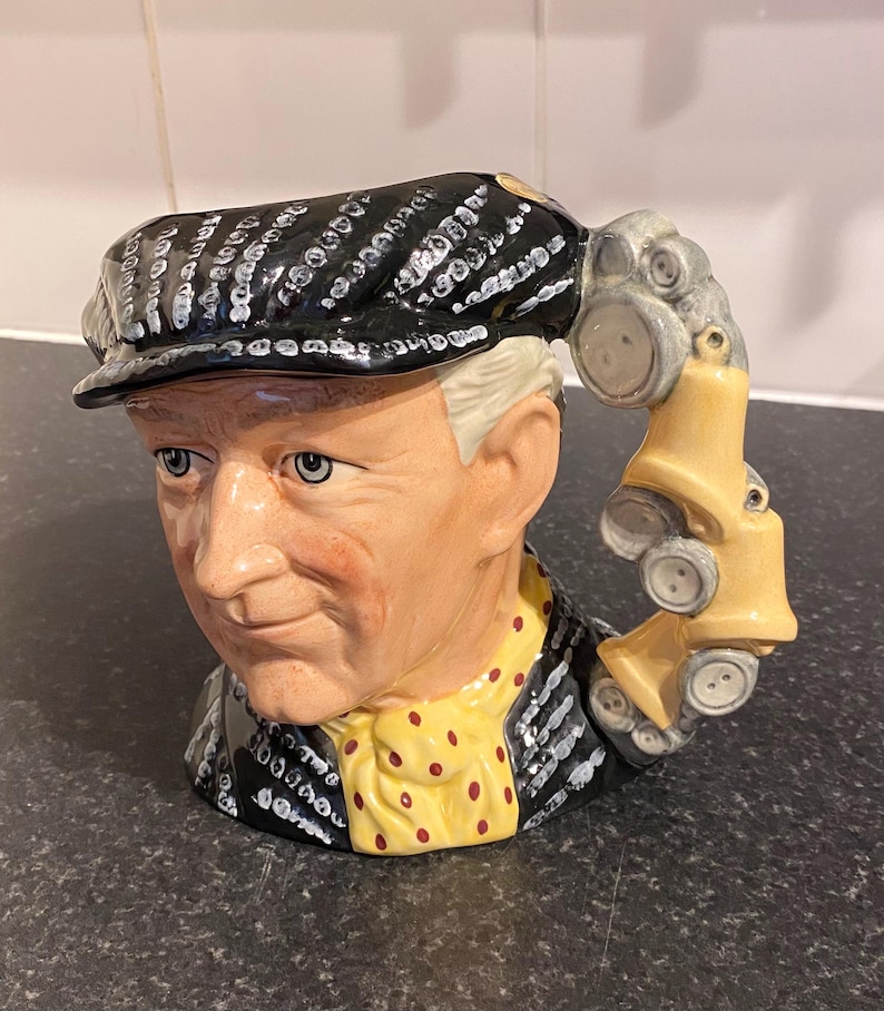 May include: A ceramic mug shaped like a man's head wearing a black and white patterned cap. The mug has a yellow and red polka dot scarf and a handle shaped like a car.
