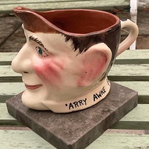 May include: A ceramic pitcher shaped like a man's head with a brown and white painted design. The pitcher has a spout and a handle. The text "'Arry Awke" is written on the bottom of the pitcher.