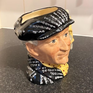 May include: A ceramic mug shaped like a man's head wearing a black hat with white polka dots. The mug has a yellow and red patterned scarf and a yellow and red polka dot tie.