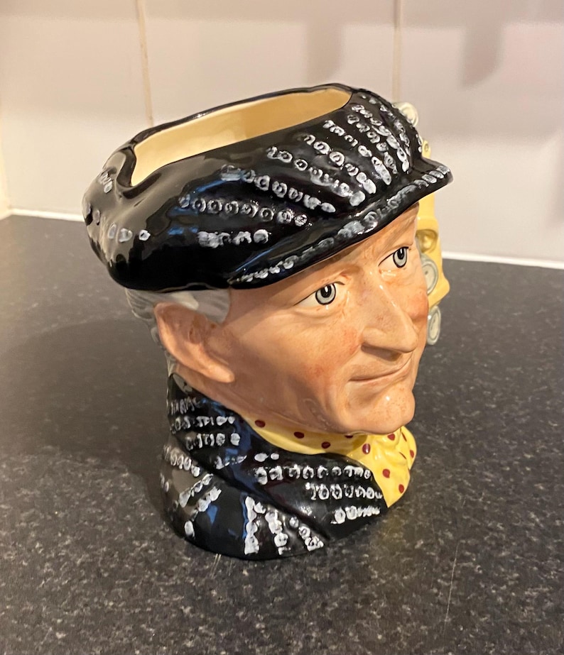 May include: A ceramic mug shaped like a man's head wearing a black hat with white polka dots. The mug has a yellow and red patterned scarf and a yellow and red polka dot tie.