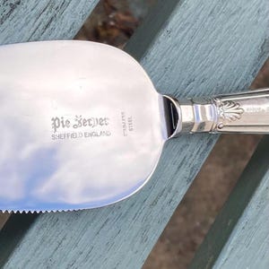 Puede incluir: Un servidor de pastel de acero inoxidable con borde dentado y mango ornamentado. Las palabras "Pie Server" y "Sheffield England" están grabadas en la hoja. El servidor es reflectante.