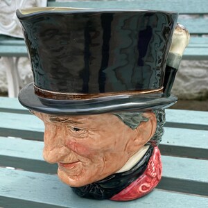May include: A novelty ceramic mug in the shape of a man's head wearing a top hat. The mug has a black top hat, a brown brim, and a cane handle. The man's coat is red and black, with a detailed face.