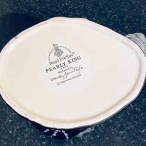 May include: White ceramic figurine of a man wearing a black hat and coat. The figurine is a Royal Doulton Pearly King, modeled by Stanley Jament, and is marked with the number D 6760. The figurine is made in 1986.
