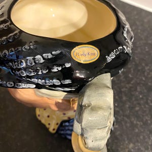 May include: A black and white ceramic pitcher with a woman's face and a black and white patterned hat. The pitcher has a gold label that reads "Pearly King".