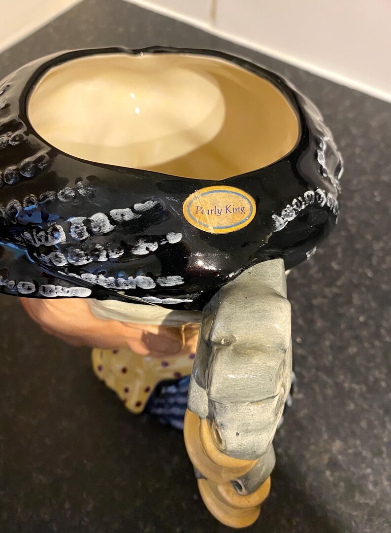 May include: A black and white ceramic pitcher with a woman's face and a black and white patterned hat. The pitcher has a gold label that reads "Pearly King".
