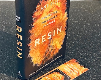 Puede incluir: Un libro titulado "Resin" de Ane Riel. La portada presenta un diseño abstracto naranja y amarillo ardiente con el título en letras negras. El libro es una historia protegida, atesorada y atrapada. Es un best-seller internacional galardonado.