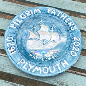 May include: A round, blue decorative plate with white raised lettering. The plate features the text "PILGRIM FATHERS 1620 2020 PLYMOUTH" and an image of a ship labeled "MAYFLOWER". The plate is on a light blue surface.