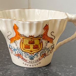 Peut inclure: Une tasse à thé en porcelaine couleur crème avec un bord cannelé et une bordure dorée. L'avant présente un blason héraldique avec deux léopards orange entourant un écu jaune contenant trois châteaux rouges, avec le texte BON ACCORD ABERDEEN.
