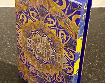 Puede incluir: Un libro titulado "The Asplin Box" con una cubierta azul y dorada con un patrón repetitivo de cisnes y serpientes. El texto "It had me spellbound" está impreso en la cubierta.