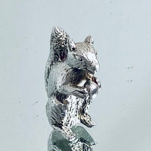May include: A silver squirrel figurine holding a nut in its paws. The squirrel is sitting upright with its tail curled up behind it.