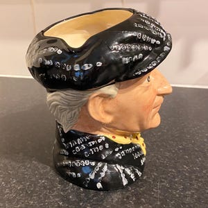 May include: A black and white ceramic mug shaped like a person's head wearing a black hat. The mug has a white pattern of dots and lines on the hat and the person's clothing.