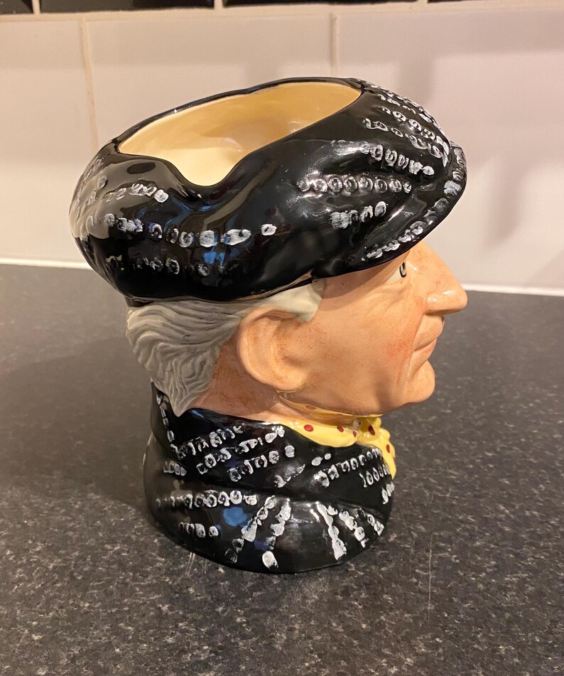 May include: A black and white ceramic mug shaped like a person's head wearing a black hat. The mug has a white pattern of dots and lines on the hat and the person's clothing.