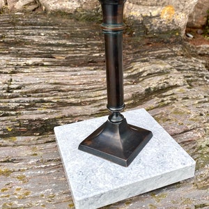 May include: A black metal candlestick with a square base and a simple design. The candlestick is sitting on a white marble base.