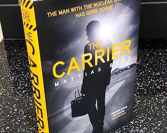 Puede incluir: Un libro de bolsillo negro y amarillo titulado "The Carrier" de Mattias Berg. La portada presenta la silueta de una persona que lleva un maletín y el texto "The Man with the Nuclear Briefcase Has Gone Rogue".
