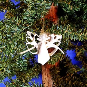 May include: A white angel Christmas ornament with a pearl head, gold halo, and ribbon accents. The angel is hanging from a gold string against a backdrop of a green Christmas tree and a brown tree trunk.